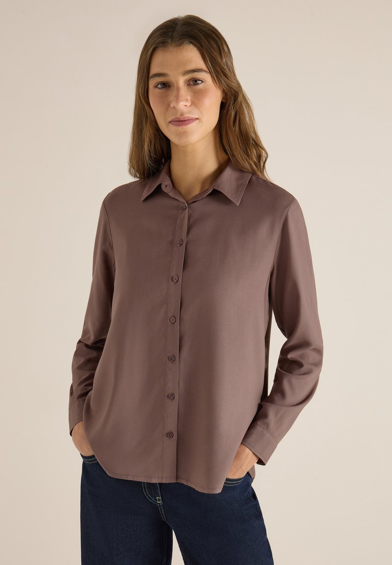 Femme aux longs cheveux bruns portant une chemise mauve à manches longues boutonnée et un jean bleu foncé, les mains dans les poches, sur un fond uni.