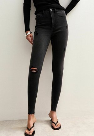 Persoon die strakke zwarte gescheurde spijkerbroek, een zwart shirt met lange mouwen, zilveren armbanden en zwarte sandalen met hakken draagt en tegen een effen muur staat.