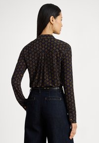 Sieviete ar gariem melniem matiem, melnu kreklu ar oranžām sēņu rakstiem, tumšām džinsu biksēm un zelta riņķveida auskarus, vērsta prom.