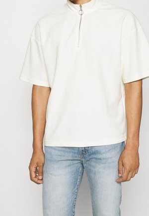 Person trägt ein weißes kurzärmeliges Oberteil mit Vorderreißverschluss und hellblaue Jeans, steht vor einem einfarbigen Hintergrund.