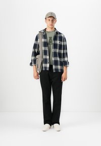 Checked flannel shirt in navy and white, olive green t-shirt with large letter, black pants, beige shoes, grey cap, and shoulder bag.