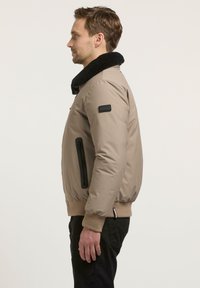 Beige bomberjakke med svart kunstpelskrage, glidelåslommer på sidene og ribbestrikkede mansjetter. Glatt tekstur med en avslappet passform.