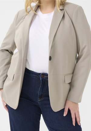 Femme portant un blazer beige sur un t-shirt blanc et un jean taille haute bleu foncé, debout avec une main sur la hanche.