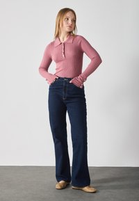 Rosa ribbet langermet topp med krage og knappdetaljer, kombinert med mørkeblå utsvingte jeans og beige ballettsko på et grått gulv.