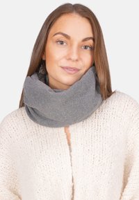 Jeune femme aux cheveux bruns lisses portant une épaisse écharpe grise et un pull en tricot crème sur un fond uni.