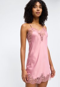 Γυναίκα που φοράει ροζ σατέν φόρεμα με τιράντες, διακοσμημένο με φλοράλ δαντέλα στο ντεκολτέ και το τελείωμα, στέκεται μπροστά σε απλό φόντο.