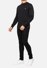 Sweatshirt noir à manches longues et col rond, avec une poche zippée sur le bras gauche et un petit logo sur la poitrine. Porté avec un jean noir et des baskets blanches.