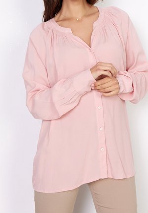Femme portant un chemisier rose ample à manches longues avec des boutons et un pantalon beige, ajustant une manche avec les deux mains.
