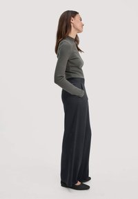 Aansluitend donkergroen langemouwen top gecombineerd met wijde zwarte broek, beide met een gestructureerde, gladde afwerking, aangevuld met zwarte platte schoenen.