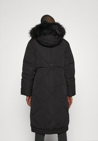 Černý plněný kabát s velkou kapucí lemovanou kožešinou, prošívaný design, dlouhé rukávy a žebrované manžety. Obsahuje stahovací pas a logo na zadní straně.