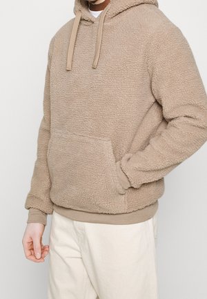 Beiger flauschiger Kapuzenpullover mit Fronttasche, getragen von einer Person mit einer Hand in der Tasche und hellen Hosen.