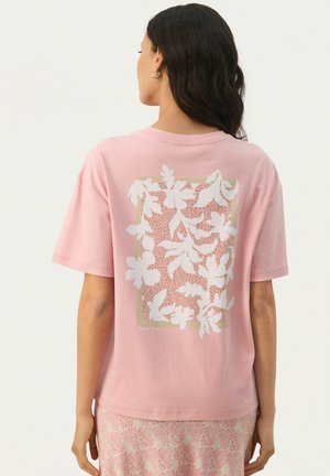 T-shirt en coton rose avec un imprimé floral blanc et une bordure rectangulaire verte claire au dos. Manches courtes et coupe décontractée.