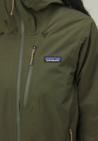 Zelena vodootporna jakna s kapuljačom, s patentnim zatvaračem na džepovima i logotipom Patagonia. Uključuje pletene detalje s trakama na kapuljači.