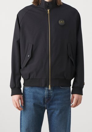 Osoba ubrana w czarną kurtkę na zamek z dwoma kieszeniami na guziki z przodu i logo EA7 na lewej piersi, do tego niebieskie dżinsy.