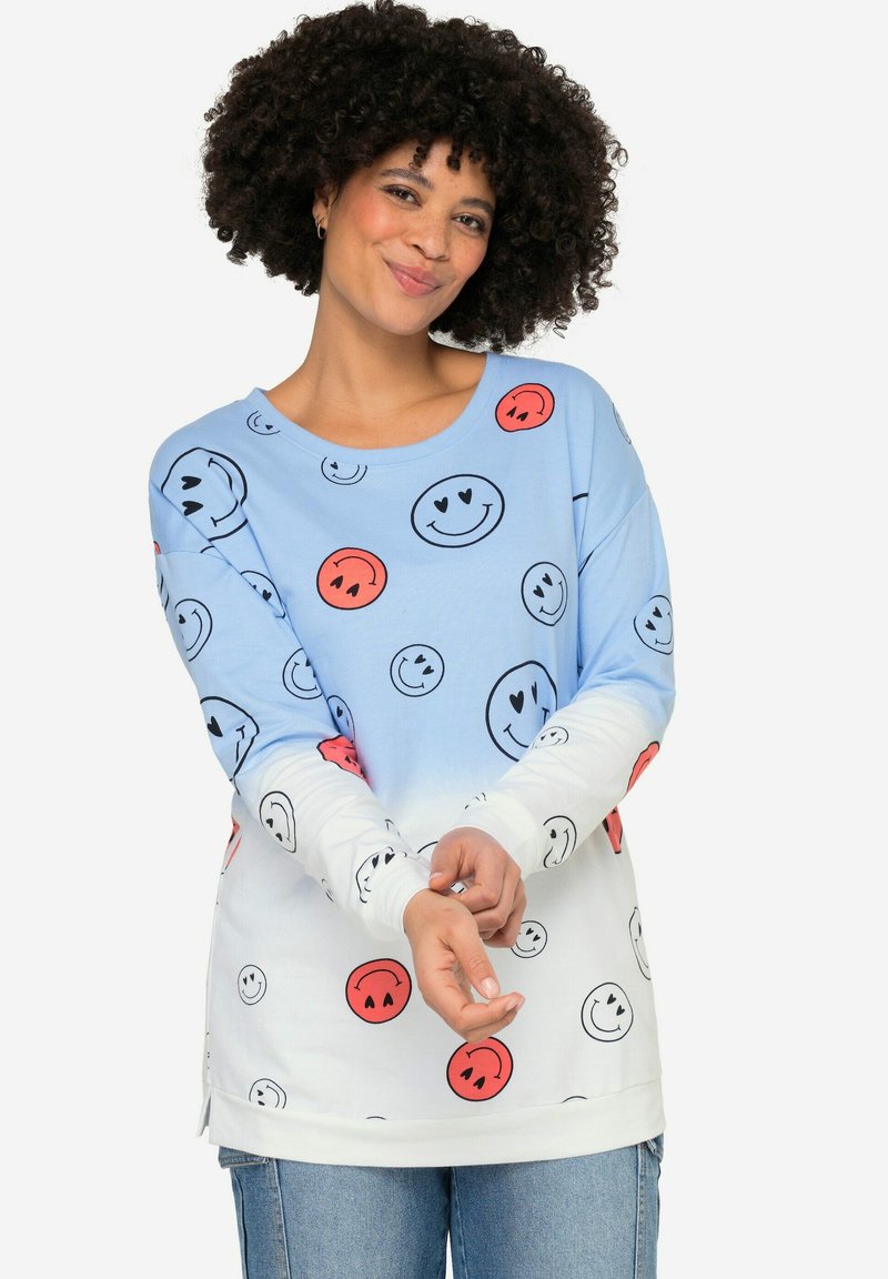 Sudadera de manga larga en azul claro y blanco, con un patrón de caras sonrientes en negro y rojo. Tejido suave, corte relajado, aberturas laterales.