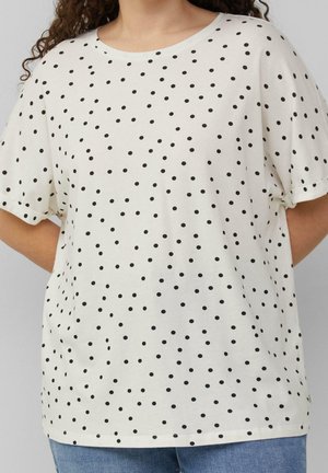 Chemise blanche à manches courtes, ample, avec de petits pois noirs dispersés, portée avec un jean bleu, les bras de la personne dans le dos.