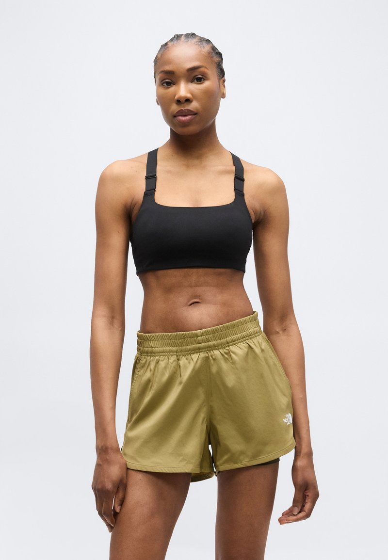 Αθλητική γυναίκα με πλεξούδες, που φοράει μαύρο αθλητικό τοπ και σορτς The North Face σε ελιά πράσινο, στέκεται μπροστά σε απλό ανοιχτόχρωμο φόντο.