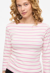 Γυναίκα με σγουρά ξανθά μαλλιά που φοράει λευκή μπλούζα με μακριά μανίκια και οριζόντιες ροζ ρίγες, κοιτάζοντας μπροστά με ουδέτερη έκφραση.