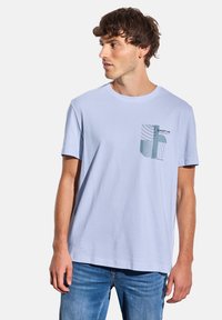 T-shirt en coton léger bleu clair avec un col rond et des manches courtes, présentant un motif graphique sur la poche imprimée en vert et noir.