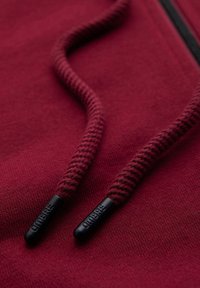 Gros plan des cordons d’une capuche bordeaux avec embouts en plastique noir embossés du mot « OMBRE », sur un tissu assorti.