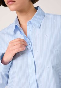 Light blue striped button-up shirt made of smooth fabric, featuring a collar and decorative buttons, with a relaxed fit and long sleeves.