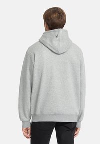Felpa grigia in cotone con cappuccio ampio, polsini e orlo a costine. Presenta un piccolo logo di ragno sulla parte posteriore. Indossata con pantaloni neri.
