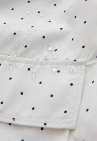 Tissu blanc à pois noirs montrant une poche cousue et des gouttes d'eau perlées à la surface, indiquant une résistance à l'eau.