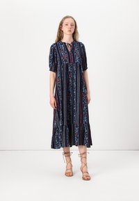Navy floral maxi dress with short puff sleeves, featuring vertical stripes, a tie neckline, and a ruffled hem. Paired with brown strappy sandals.