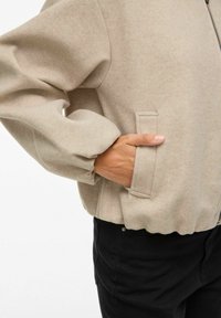 Beige flīsa jaka ar rāvējslēdzēju priekšpusē, pārmērīgiem piedurknēm un sānu kabatu. Iezīmēta ar viļņotu apakšmalu un gludu tekstūru. Savienota ar melniem biksēm.