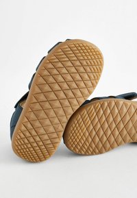 Blaue Slipper mit einer strukturierten Gummisohle, die ein geometrisches Muster aufweist, sowie einem Obermaterial aus weichem Stoff mit genähten Details.