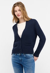 Marinblå stickad cardigan med V-hals, framknappar och två påsydda fickor. Har ribbade ärmslut och nederkant för en texturerad finish.