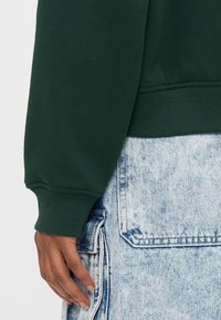 Grüner Pullover mit gerippten Bündchen an Ärmeln und Saum, kombiniert mit einem hellblauen Jeansrock mit großen Taschen und verwaschener Optik.