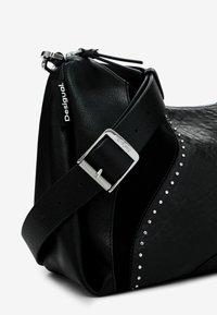 Zwarte leren handtas met een gestructureerde afwerking, zilveren studs aan één zijde, een verstelbare schouderband met een metalen gesp en een ritsluiting.