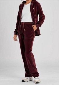 Personne portant un sweat à capuche en velours bordeaux avec fermeture éclair et un pantalon assorti, portant un T-shirt blanc et des baskets blanches, se tenant devant un fond uni.