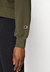 Olivgrüner Sweatshirt mit gerippten Bündchen, das ein kleines gesticktes Logo in Rot, Weiß und Schwarz am linken Ärmel aufweist. Schwarzer Stoff darunter.