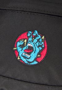 Pache de tecido preto com um design bordado de uma mão azul com dentes afiados dentro de um círculo vermelho, detalhado com acentos amarelos.