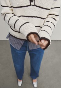 Pull crème tricoté avec rayures noires, col boutonné et poignets côtelés, porté sur une chemise rayée bleu et blanc ; jean large bleu et chaussures argentées.
