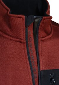 Rote Fleecejacke mit hohem Kragen, schwarzen Akzenten und einem Reißverschluss vorne. Verfügt über strukturierten Stoff und ein Logo im Bereich des Reißverschlusses.