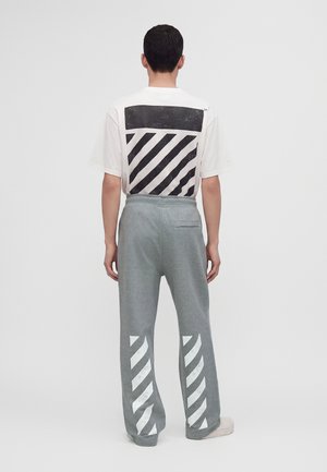 Mann trägt weißes T-Shirt mit schwarzen diagonalen Streifen auf dem Rücken und graue Hose mit weißen diagonalen Streifen an den unteren Beinen, steht mit dem Rücken zugewandt.