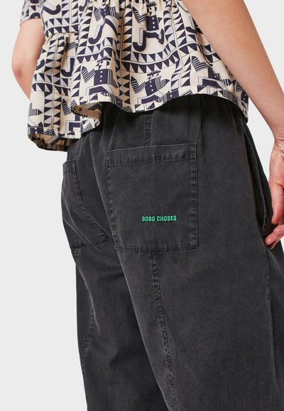 Women's outfit featuring a patterned beige top with geometric designs and black corduroy pants with a back pocket displaying green embroidery.