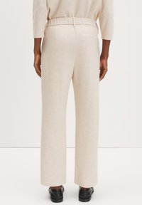 Beige wijde broek van zacht materiaal met een elastische tailleband, voorzien van subtiele textuur en zijzakken. Zwarte schoenen worden gedragen.