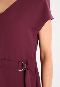 Burgunderfarbenes Kleid mit V-Ausschnitt, kurzen Ärmeln und einem Stoffgürtel mit einer silbernen runden Schnalle in der Taille für Struktur. Glatte Textur.