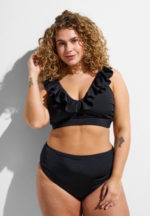 Curly-haired woman wearing black ruffled bikini top and high-waisted bottoms, standing against a white wall with tattoos on both arms.