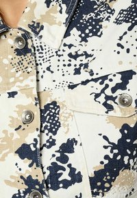 Tejido de patrón beige y azul marino, que presenta una mezcla de formas abstractas y texturas. Incluye detalles en botón plateado y bolsillos.