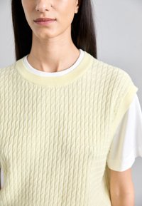Világos sárga textúrázott kötött felső, amely egy fehér pólóra van rétegezve, bordázott nyakkivágással és vállon végződő ujjakkal, struktúrált sziluettet alkotva.