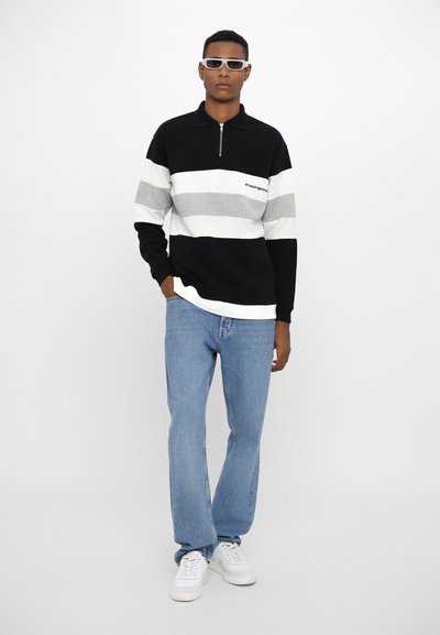 Zwart, grijs en witte gestreepte pullover met ritssluiting, gecombineerd met lichtblauwe jeans en witte sneakers. Accessoires zijn onder andere witte zonnebril.