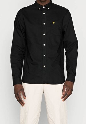 Uomo che indossa una camicia nera con bottoni con un logo di uccello giallo sulla tasca del petto e pantaloni bianchi su sfondo chiaro.