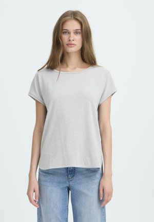 Junge Frau mit langen braunen Haaren, die ein hellgraues Kurzarm-T-Shirt und blaue Jeans trägt, steht vor einem schlichten weißen Hintergrund.