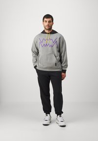 Sudadera gris con diseño de zigzag morado, capucha negra y cordón amarillo; combinada con pantalones de chándal negros y zapatillas blancas.