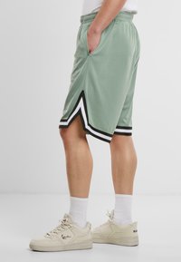 Lys grønne træningsshorts med sorte og hvide side accenter, elastisk talje og lommer. Båret med beige sneakers og hvide sokker.